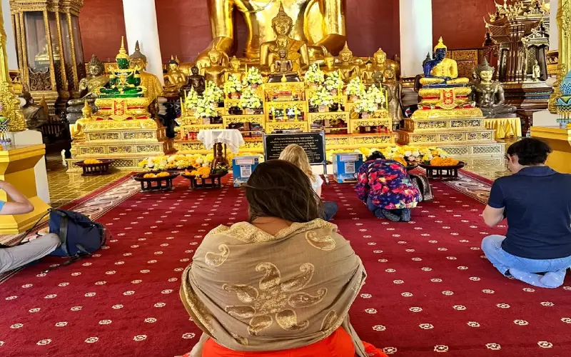 Fiéis em oração diante do Buddha Sihing, envoltos na serenidade do Wat Phra Singh. (Crédito: @thaisviagemtur)